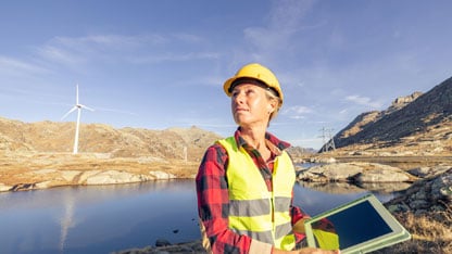 Un ingeniero está junto a una turbina eólica en la orilla de un lago sosteniendo una tableta digital.