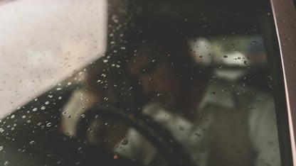 Un hombre dentro de un automóvil visto a través de un parabrisas con gotas de lluvia.